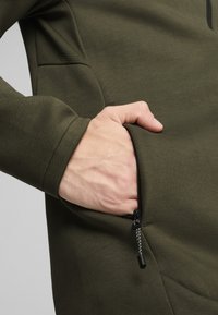 Donkergroen pullover met een gestructureerd schouderpaneel, een ritszak en een hand die in de zak is gestoken, wat de gladde textuur van de stof laat zien.