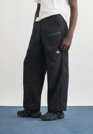 Person iført løstsittende svarte North Face-bukser med glidelåslommer og svarte slip-on sko, stående på blått teppe.