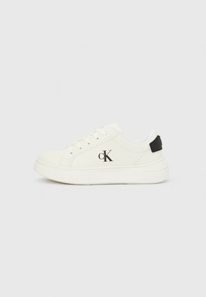Zapatilla blanca de perfil bajo con diseño de cordones, logo "cK" negro en el lateral y parche negro en el talón sobre un fondo claro y liso.