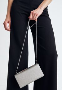 Femme tenant une pochette rectangulaire en argent par une chaîne, debout dans un pantalon large noir contre un fond uni.
