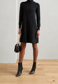 Abito nero a maglia con maniche lunghe e collo alto, abbinato a stivaletti neri alla caviglia e una piccola borsa nera con tracolla in oro.