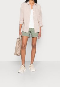 Lysbeige linned skjorte med knapper over en hvid V-udskæring top, parret med olivengrønne shorts og beige sneakers. Holder en beige tote bag.
