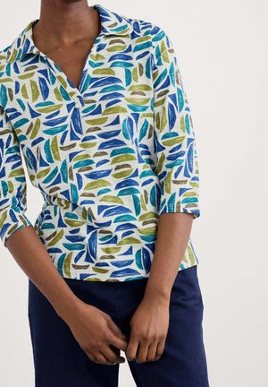 Persoon draagt een overhemd met knopen met blauwe, groene en bruine abstracte bladmotieven en een donkerblauwe broek, handen in elkaar gevouwen voor zich.