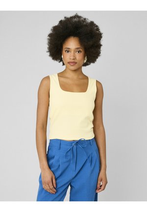 Femme avec une coiffure afro naturelle portant un haut sans manches jaune pâle et un pantalon bleu taille haute avec une ceinture nouée, debout devant un fond gris.