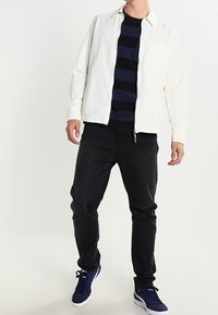 Herretøj bestående af en lys cremet lynlåsjakke, en marineblå stribet t-shirt, sorte jeans og marineblå sneakers med hvide såler.