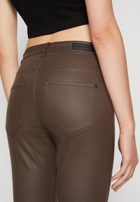 Brune, tettsittende jeans har en jevn tekstur, baklommer med en spiss design, og en svart preget merkevarelabel på livet.