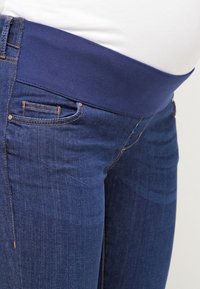 Mörkblå gravidjeans med bred elastisk midja, bärs med en vit topp, visar framfickor och sömdetaljer.