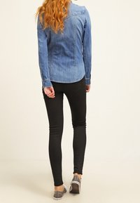 Vrouw met golvend rood haar, gekleed in een blauw denim shirt, zwarte skinny broek en grijze sneakers, die wegloopt op een effen lichte achtergrond.