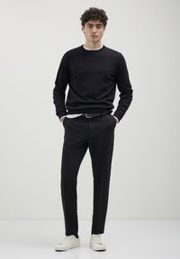 Uomo in piedi con le mani in tasca, indossa un maglione nero, pantaloni neri, scarpe bianche e una cintura, su uno sfondo grigio uniforme.