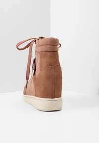 Hnědý semišový kotníkový tenisky s klínovým podpatkem, polstrovaným límcem, šněrováním vpředu, bočním zipem a bílou gumovou podrážkou, zobrazený zezadu.