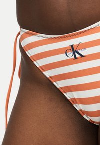 Lähivaade inimese puusast, kes kannab oranži- ja valge triibulise mustriga Calvin Kleini bikiinipükse, millel on külgtiepaelad ja nähtav õmblus.