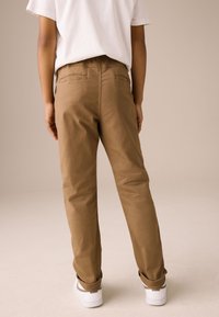 Pantaloni maro din bumbac, cu croială lejeră, având buzunare la spate și tivuri îndesate, asortați cu adidași albi pe un fundal neutru.