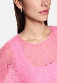 Rosa gestrickter Pullover mit einem strukturierten, offenen Design und einem weiten Ausschnitt, kombiniert mit verdrehten silbernen Creolen.
