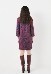 Bordeauxrotes Kleid mit einem marineblauen Paisleymuster, dreiviertellangen Ärmeln und einem ausgestellten Saum, kombiniert mit kniehohen braunen Lederstiefeln.