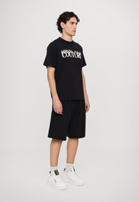 Juodas marškinėlis su "VERSACE JEANS COUTURE" logotipu baltos ir auksinės spalvos, derinamas su juodomis šortais ir baltomis sportsbatėmis. Kasdienis stilius.