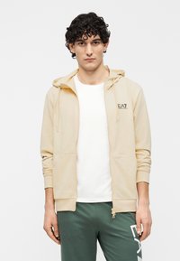 Lichtbeige hoodie met rits en trekkoord aan de capuchon, twee zakken aan de voorkant en geribbelde boorden. Gecombineerd met een wit T-shirt en groene joggingbroek.