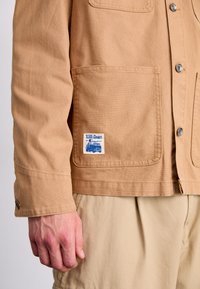 Beige kanvasjakke med knappestengning, brystlomme og merkeetikett. Har rett snitt og mansjett detaljer. Båret over lyse bukser.