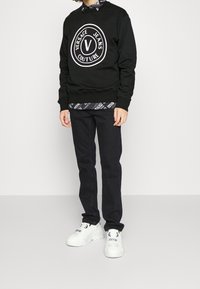 Svart sweatshirt med vit cirkulär logotyp, mönstrad skjorta under, mörka denimjeans och vita tjocka sneakers.