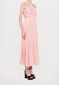 Robe maxi plissée rose avec des bretelles entrecroisées et des détails découpés. Assortie à des talons compensés noirs avec un accent en nœud.