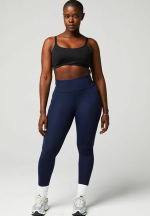 Un crop top noir associé à un legging bleu marine taille haute. Les leggings présentent une texture lisse et des détails cousus, complétés par des baskets blanches.