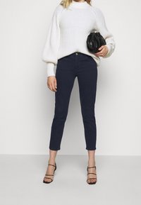 Mörkblå tajta jeans med medelhög midja, i kombination med en strukturerad vit stickad tröja och en svart clutch, iförd med svarta remsandaler.