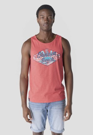 Joven con cabello corto y rizado vistiendo una camiseta sin mangas color coral con la inscripción "Okinawa Beach" y shorts de mezclilla azul claro remangados.