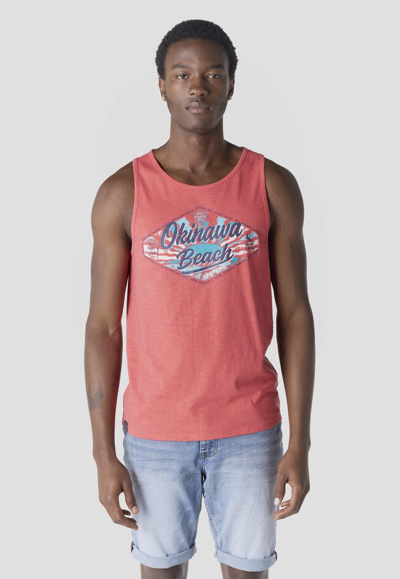 Joven con cabello corto y rizado vistiendo una camiseta sin mangas color coral con la inscripción "Okinawa Beach" y shorts de mezclilla azul claro remangados.