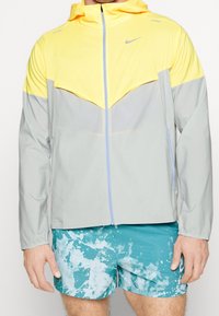 Muž v žlutošedé mikině Nike s kapucí a plným zipem a tyrkysových šortkách s vzorem.