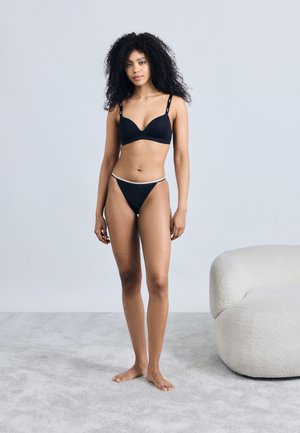 Black padded bra and matching thong with a thin waistband, displayed on a model standing on a light gray carpet, with a light backdrop.