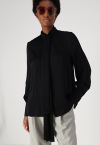 Schwarze Bluse mit langen Ärmeln und gebundenem Kragen, aus glattem Stoff gefertigt. Kombiniert mit hellgrauen, gestreiften Hosen mit lockerer Passform.