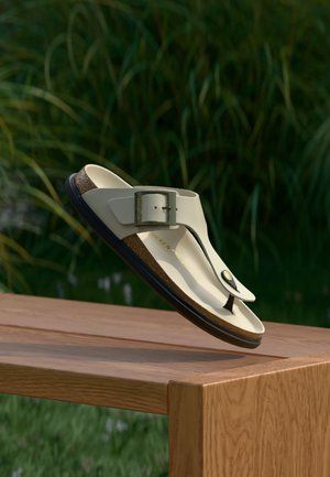 Μπεζ σανδάλι με λουράκι και ρυθμιζόμενη αγκράφα, με φελλό πάτο, να αιωρείται πάνω από ξύλινο τραπέζι με φόντο πράσινα φύλλα.