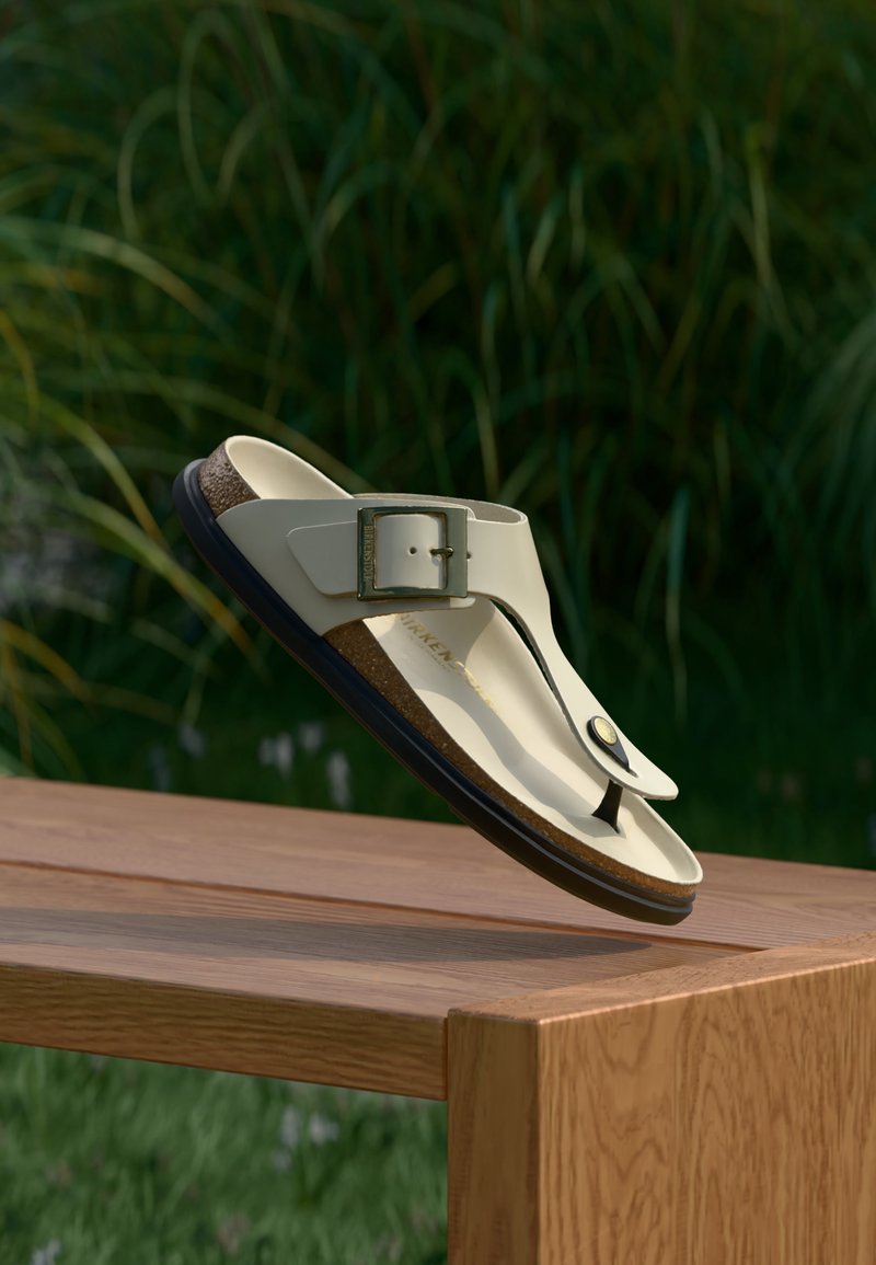 Μπεζ σανδάλι με λουράκι και ρυθμιζόμενη αγκράφα, με φελλό πάτο, να αιωρείται πάνω από ξύλινο τραπέζι με φόντο πράσινα φύλλα.