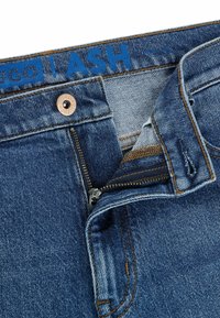 Jeans i denim med en blå etikett, kopparknapp och dragkedja. Har kontrasterande orange sömmar och en urtvättad struktur på tyget.