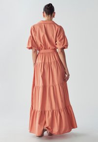 Maximeettinen mekko pehmeästä korallinvärisestä kankaasta, jossa on puffi hihat, kaulustoppi ja kerroksittain tehty röyhelöhelma, vyö vyötäröllä.