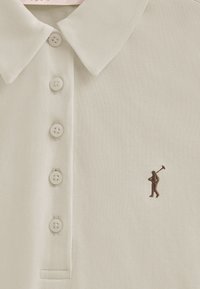 Beige poloshirt van textuurstof met een kraag, vijf knopen voor sluiting en een kleine bruine geborduurde figuur op de borst.