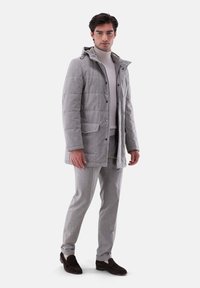 Grau gesteppte Jacke mit Kapuze, zwei Fronttaschen, kombiniert mit einem hellgrauen Rollkragenpullover und grauen Hosen, abgerundet mit dunklen Schuhen.