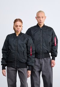 Jachete bomber negre cu un model texturat, manșete și tiv ondulate, având un fermoar în față și un etichetă din material roșu pe braț.