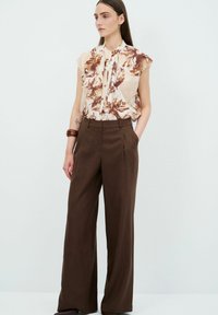 Lichte bloemige blouse met gerimpelde kraag en knopen, gecombineerd met high-waisted, wijde bruine broek, wat een ontspannen silhouet creëert.