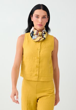 Femme aux cheveux noirs portant un haut sans manches jaune moutarde boutonné et un pantalon assorti avec une écharpe à motifs multicolores autour du cou.