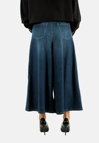 Pantalones de mezclilla anchos hasta los pies en azul oscuro, con bolsillos traseros decorativos y una textura suave. El dobladillo cae por debajo de la pantorrilla.