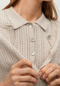 Beige neulottu neuletakki, jossa on kuvioitu pinta, kauluksellinen pääntie ja vaaleansiniset napit. Kangas on pehmeää ja hieman väljä istuvuudeltaan.