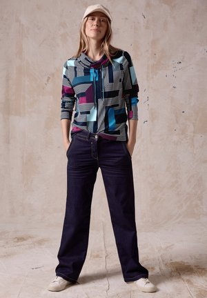 Bunt gemusterte geometrische Bluse mit navyblauen Jeans. Die Bluse hat eine Kapuze und einen lockeren Schnitt; die Hose hat einen geraden Schnitt und Taschen.