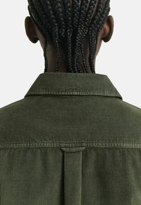 Person with black braided hair viewed from behind, wearing an olive green corduroy jacket with a back vent detail beneath the collar.