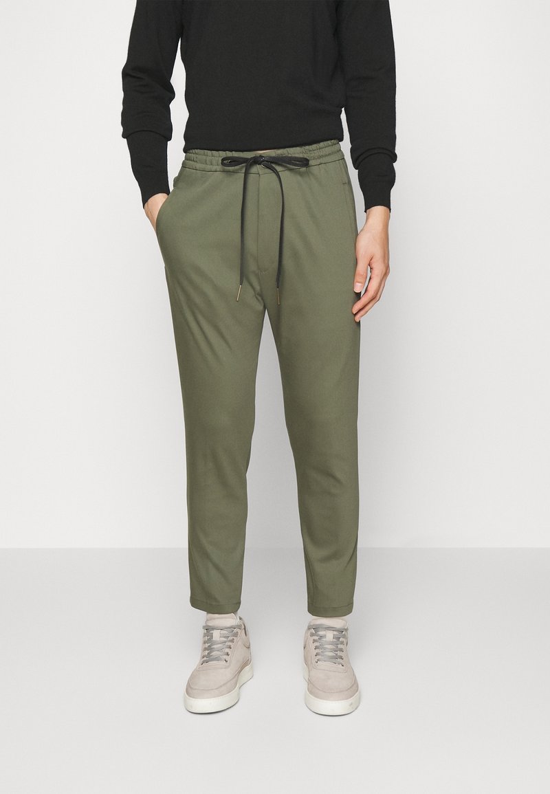 Homme portant un pantalon de jogging vert olive avec un cordon noir, un t-shirt à manches longues noir et des baskets beiges, debout les mains dans les poches.
