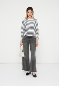 Camicia a strisce con maniche lunghe, jeans grigi chiari a gamba larga, ballerine nere e una piccola borsa bianca. Il modello è in piedi su uno sfondo neutro.