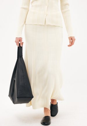 Person trägt einen cremefarbenen, gerippten Rock und ein passendes, geknöpftes Oberteil, hält eine große schwarze Ledertasche und trägt schwarze Slip-On-Schuhe.