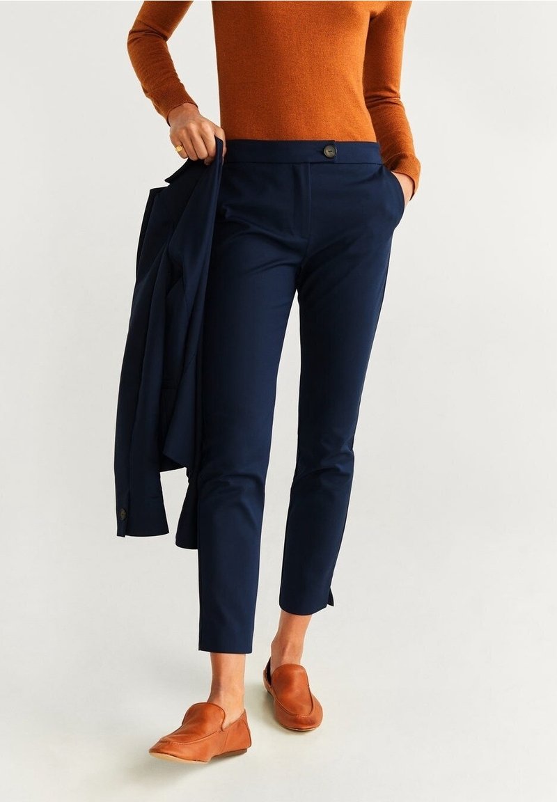 Femme portant un pantalon slim bleu marine aux chevilles, tenant un blazer bleu marine assorti, associé à un pull orange rouille et des chaussures marron sans lacets.