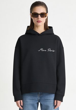 Donna che indossa una felpa nera con cappuccio con il testo "Mira Paris" sul petto, jeans blu e occhiali da sole tartarugati, in piedi davanti a uno sfondo bianco.