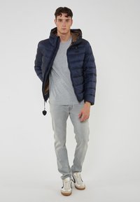 Giovane uomo che indossa un giubbotto imbottito blu navy sopra una maglietta grigia, jeans grigio chiaro e sneakers bianche, in piedi di fronte a uno sfondo uniforme.