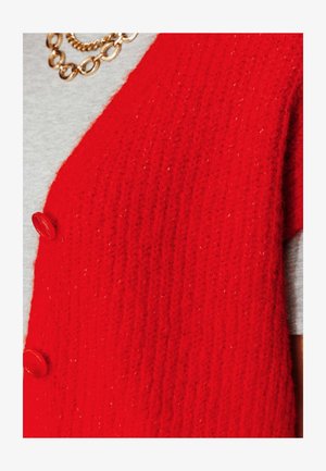 Veste en tricot rouge avec une texture côtelée et deux boutons ronds rouges. Portée sur une chemise grise, mettant en valeur sa couleur vive et son design.